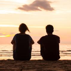 Rear view photo of two friends sitting together on beach at sunset