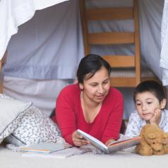 A mom reads with her son under a blanket fort.