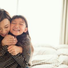 Parent and child hugging happily on bed, smiling at each other