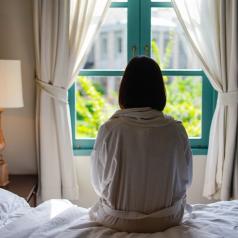 Person with short dark hair sits on bed in robe looking out open window