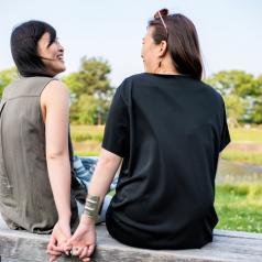 Couple sit on bench, laughing and talking openly, hands resting off the side of the bench, joined by a fingertip