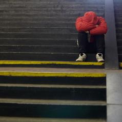 A figure in a red jacket sits on some stairs. Their face and hands are hidden.