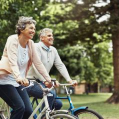 Older couple takes bike ride in tree-lined park. Both are smiling happily