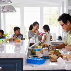 One parent feeds children breakfast while other parent makes lunches in open kitchen