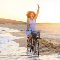 Person in sundress rides bike in waves at beach, waving at someone in distance