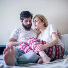 A young man and woman lean into each other as they sit on an unmade bed.