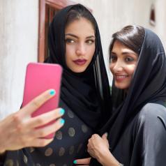 Two young women in hijabs take a selfie with a pink smartphone.