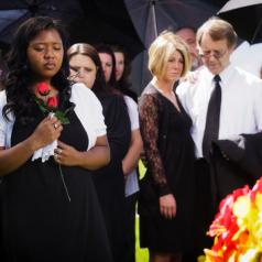 A crowd gathers to mourn at a funeral. A woman in front clutches roses to her chest.
