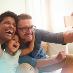 Family with young child laughs as they take a selfie with a smartphone.