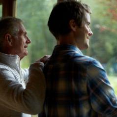 Thoughtful parent and adult son stand at window of cottage and look out toward woods
