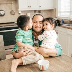Parent sits at table and hugs two happy toddlers sitting on table in tidy kitchen