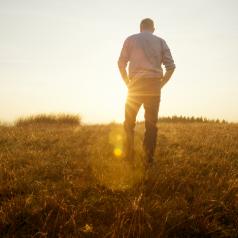 Rear view of person with short hair in pants and sweater walking in field at sunset