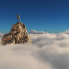 Man stands on pointed, rocky cliff that juts out above a layer of clouds.