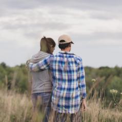Rear view photo of two adults standing in field under cloudy sky. Person in flannel shirt and baseball cap has arm around person with long ponytail