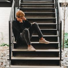 Young adult sits on stairs outside house with hands covering face looking down