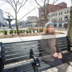 Half-transparent photo of person with curly hair wearing hat and coat sitting on bench on cold day
