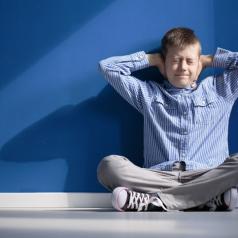 A young boy in a blue shirt sits against a blue wall, covering his ears with both hands.