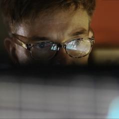 A man looks at a computer screen, which reflects in his glasses